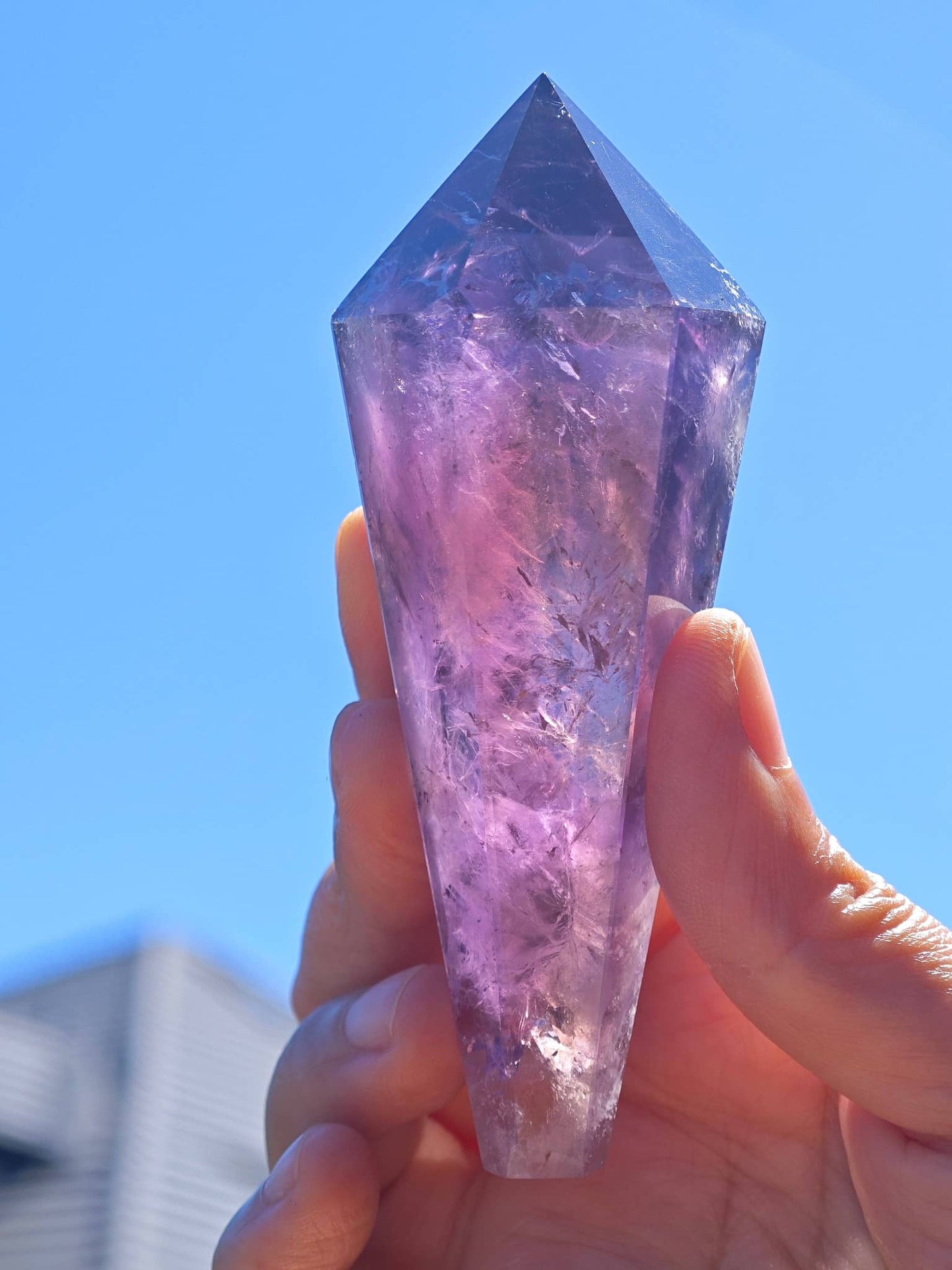 Hand holding a Natural Brazilian Amethyst Wand glimmering in sunlight against a clear blue sky