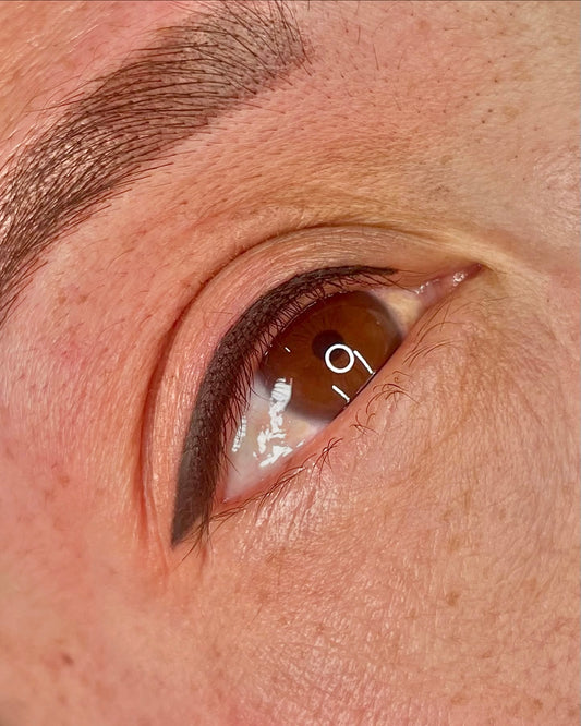 Close-up of a brown eye with black eyeliner tattoo, showing the product applied along the upper lash line, branded Black Dahlia Botánica.