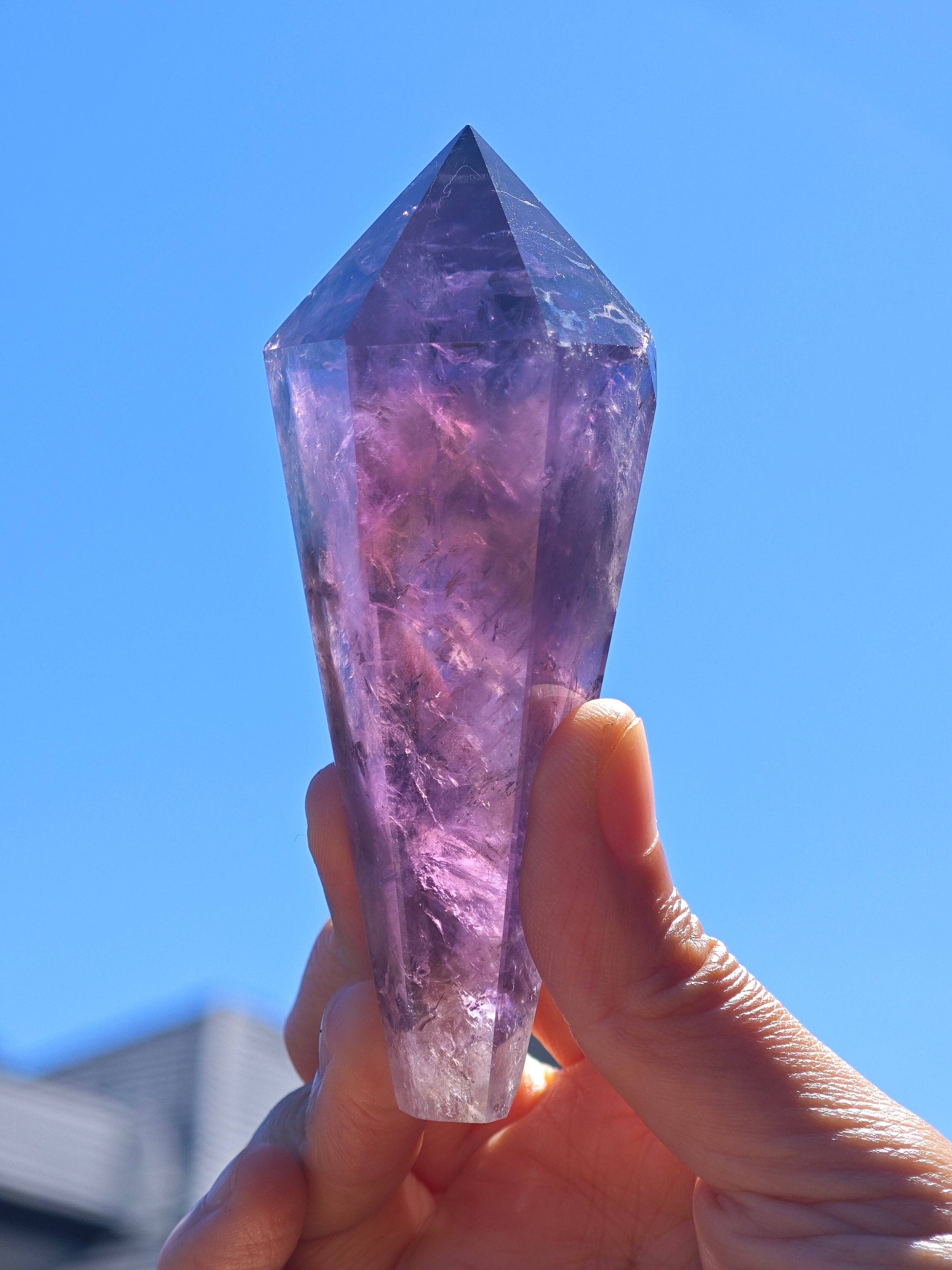 Hand holding a Natural Brazilian Amethyst Wand with a clear purple hue against a bright blue sky